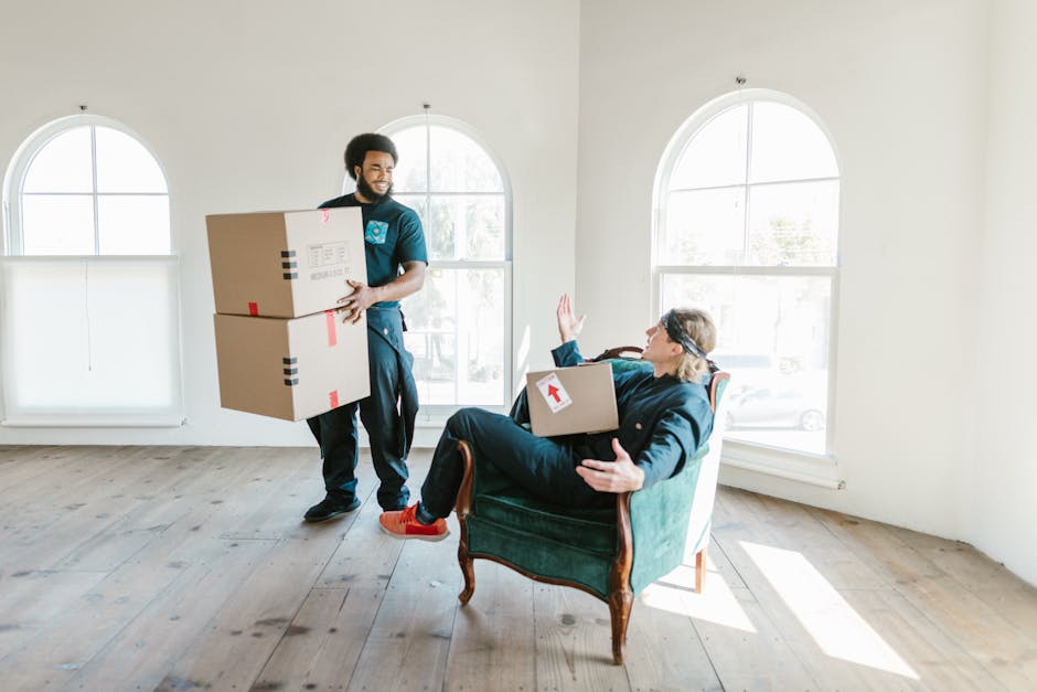 A woman with curly dark hair wearing a grey hoodie is kneeling on the floor beside a sofa, carefully wrapping it in white packing paper or protective padding as part of the home relocation process. Th